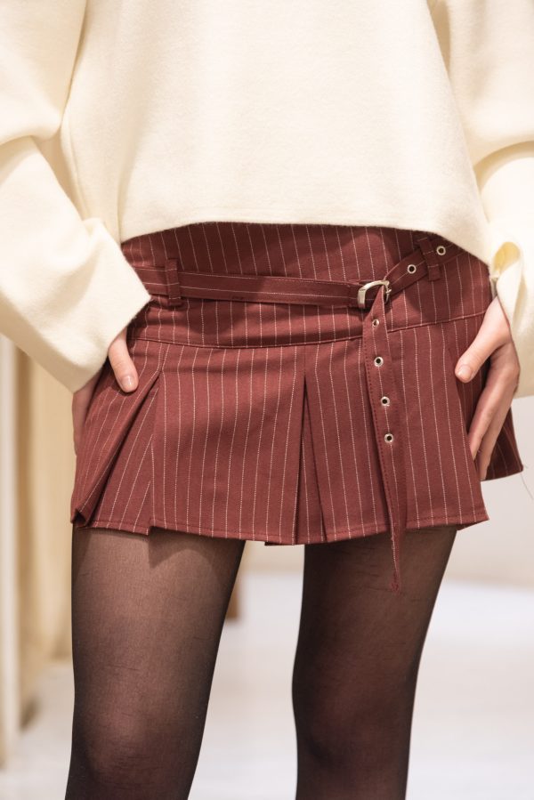 Falda mini tableada en tono bordo con finas rayas verticales blancas y detalle de cinturón, usada con un suéter crudo y medias negras.