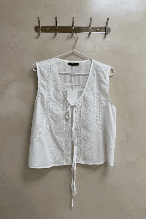 Chaleco blanco sin mangas con escote en V, adornado con puntilla y bordados florales a tono. Confeccionado en poplin 100% algodón, con cordones para anudar al frente.